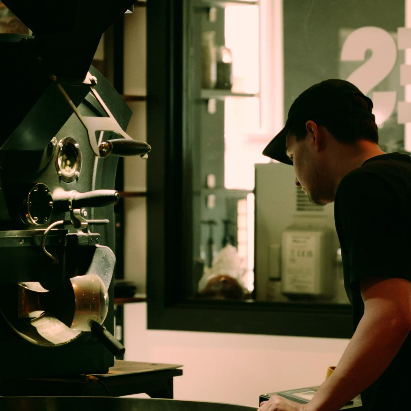 A roaster in a black cap working at the drum roaster in the Peoples Coffee roastery, with the cafe through the doorway behind